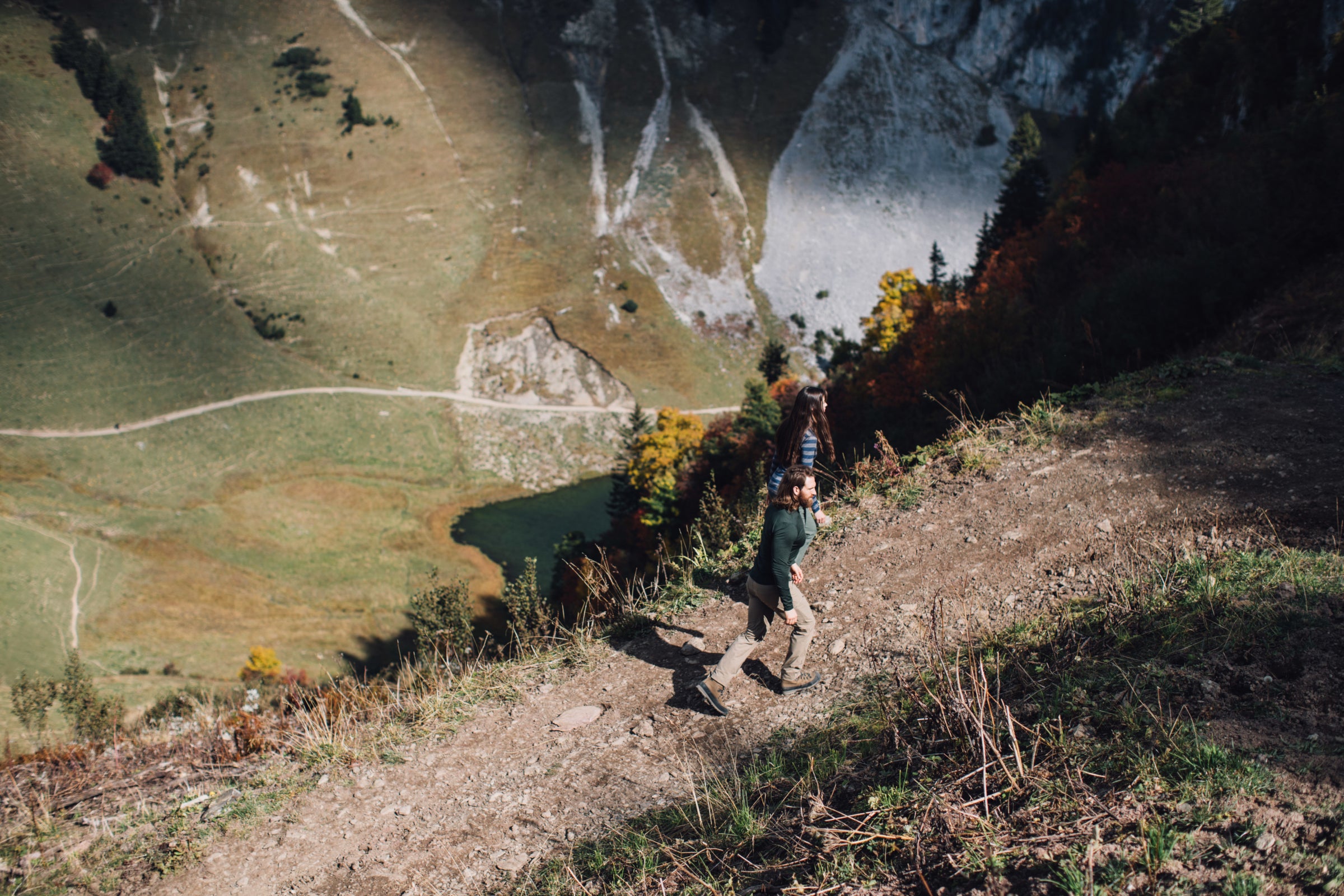 Two hikers descending a steep mountain trail with a view of a lake and trails below in a valley.