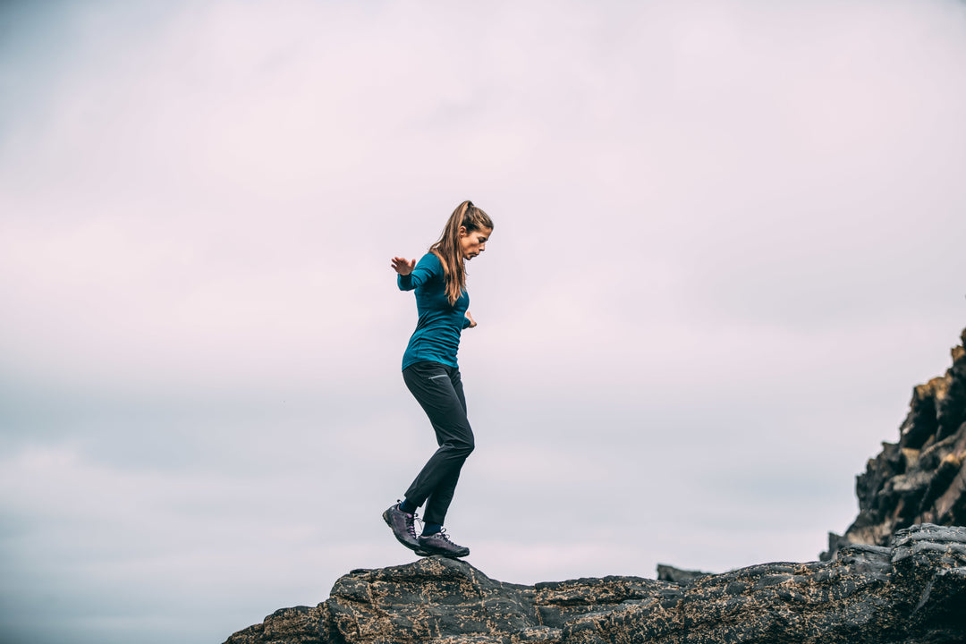 A woman in Isobaa merino clothing balances on a rocky outcrop with a pale sky in the background, focused and poised as she navigates the uneven terrain