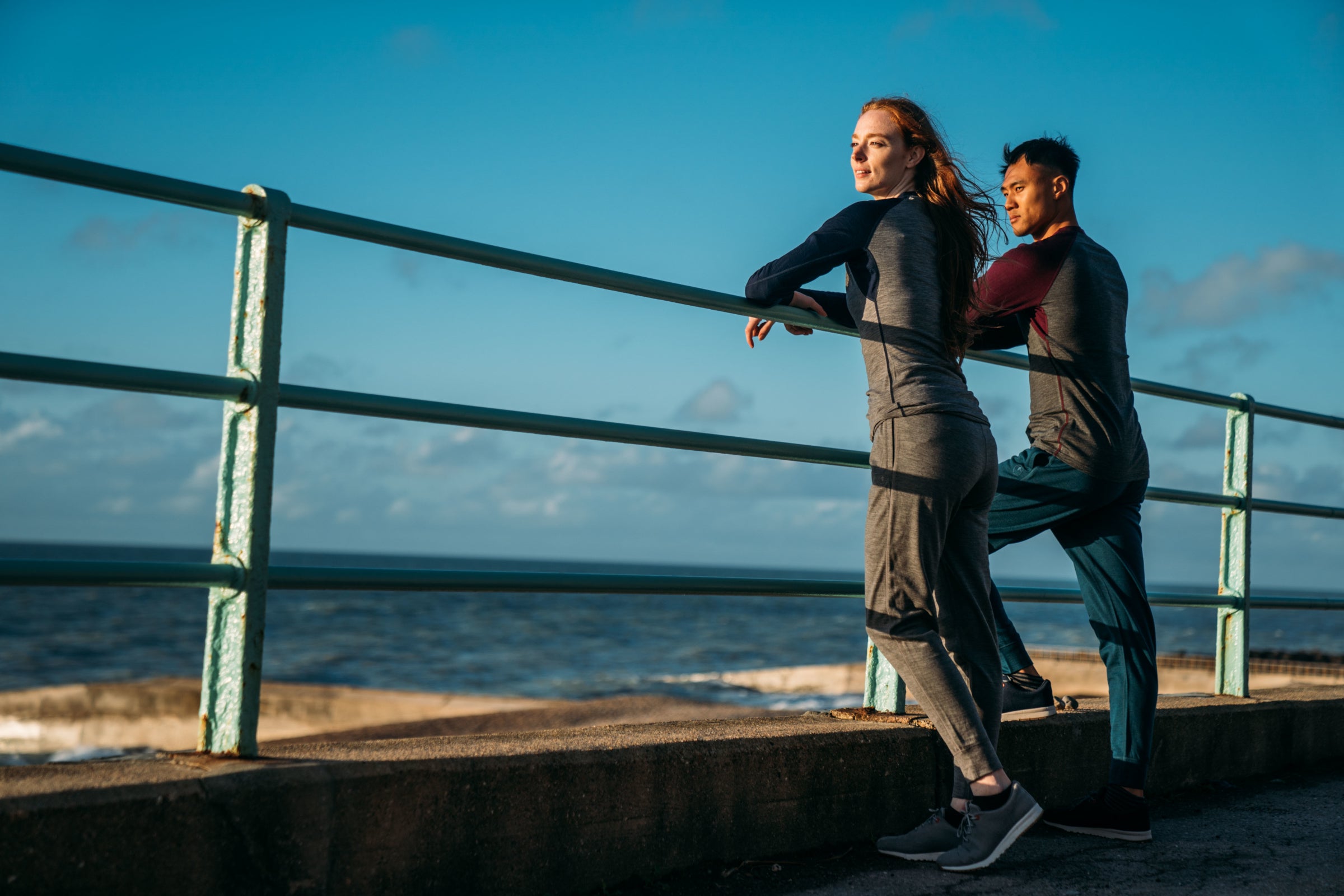 Two people wearing Isobaa merino activewear stand by a seaside railing, looking out at the ocean under a clear, blue sky