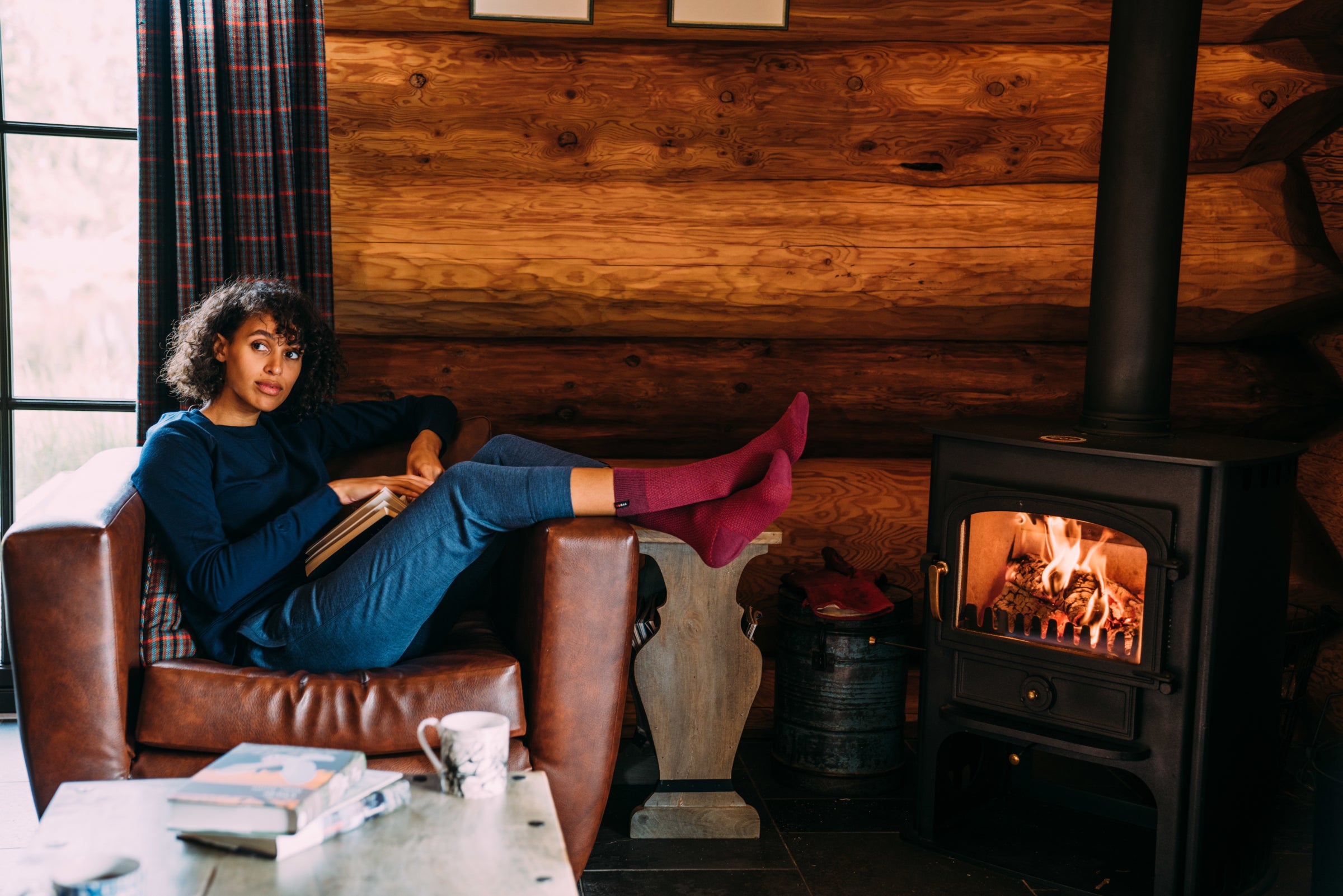 A person wearing Isobaa merino loungewear, relaxing with a book in a cosy log cabin, sitting on a leather armchair near a wood-burning stove