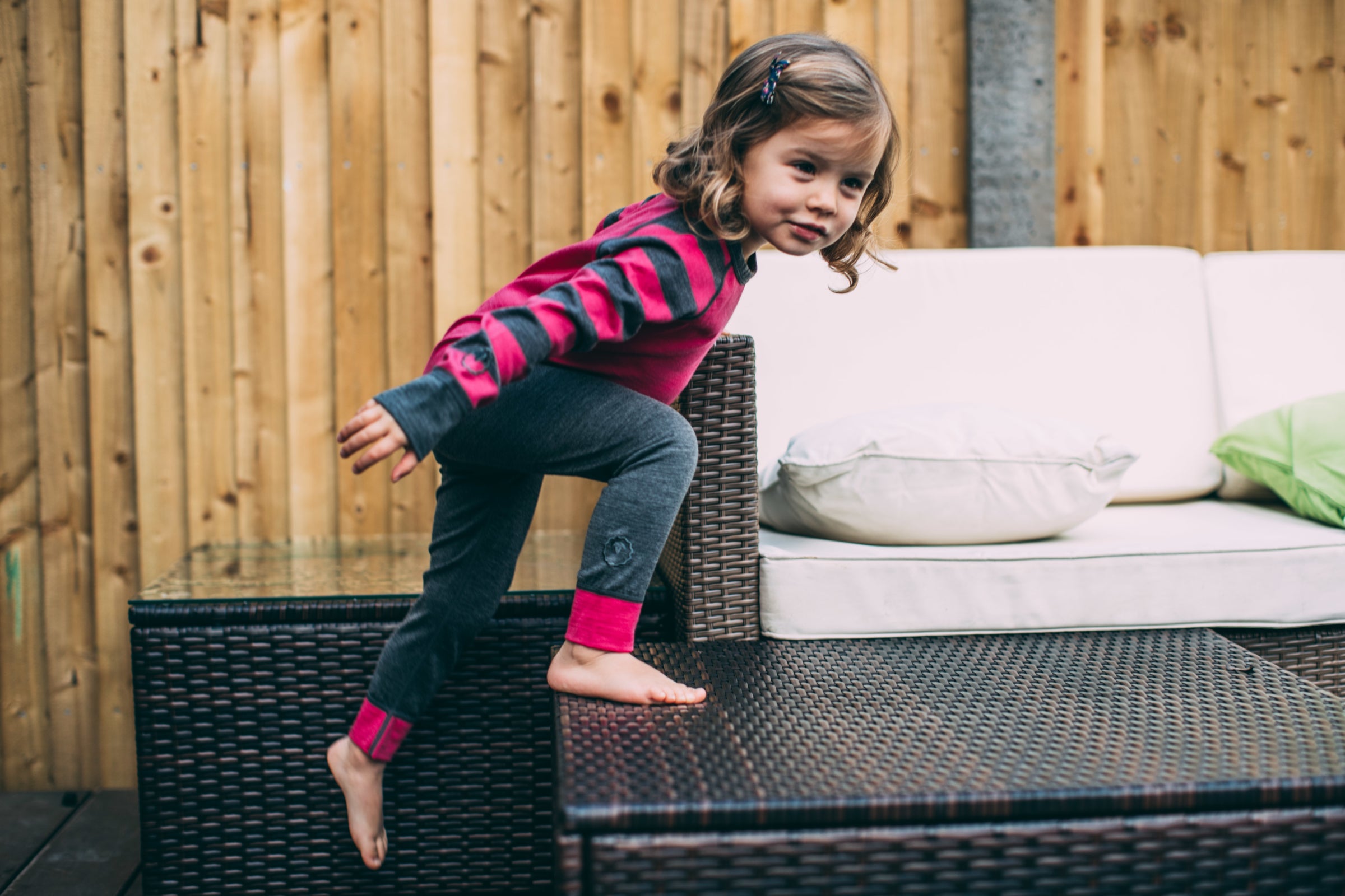 A young girl climbing on outdoor furniture, wearing a pink and grey striped Isobaa Merino Blend 200 Long Sleeve Crew and Merino Blend 200 Leggings