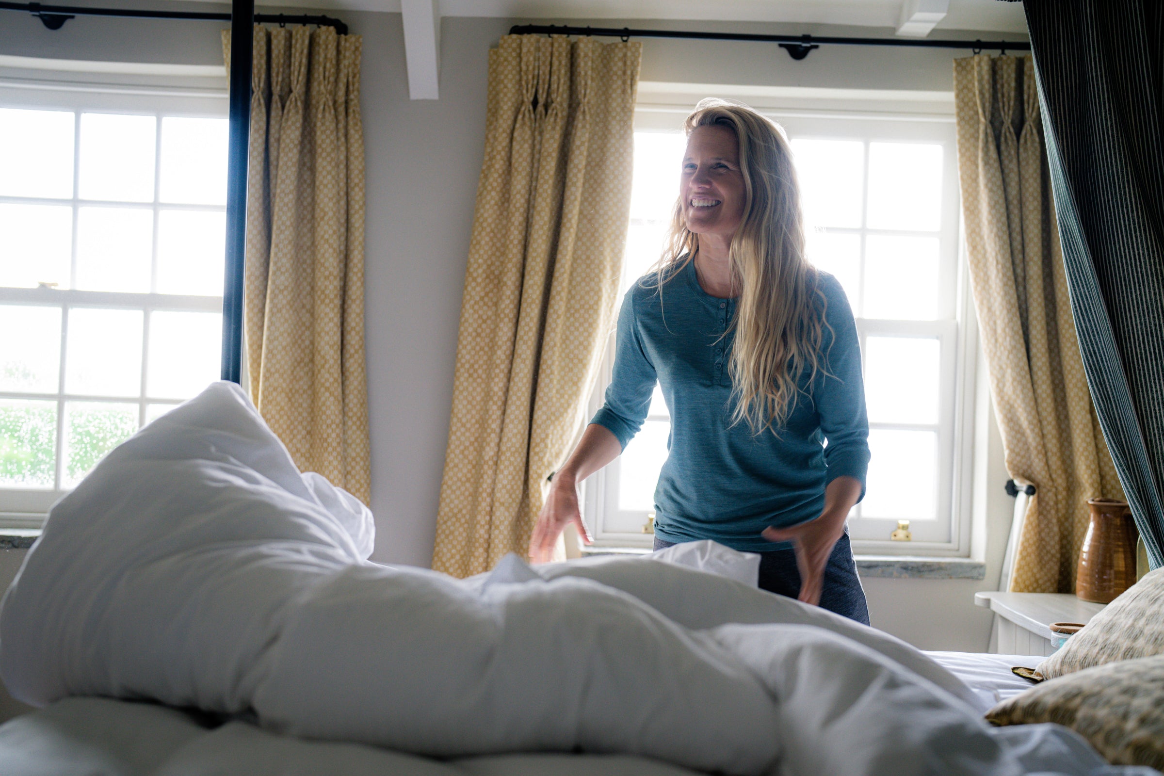 Woman smiling while making a bed, wearing Isobaa merino sleepwear in a cozy, naturally lit bedroom with yellow patterned curtains