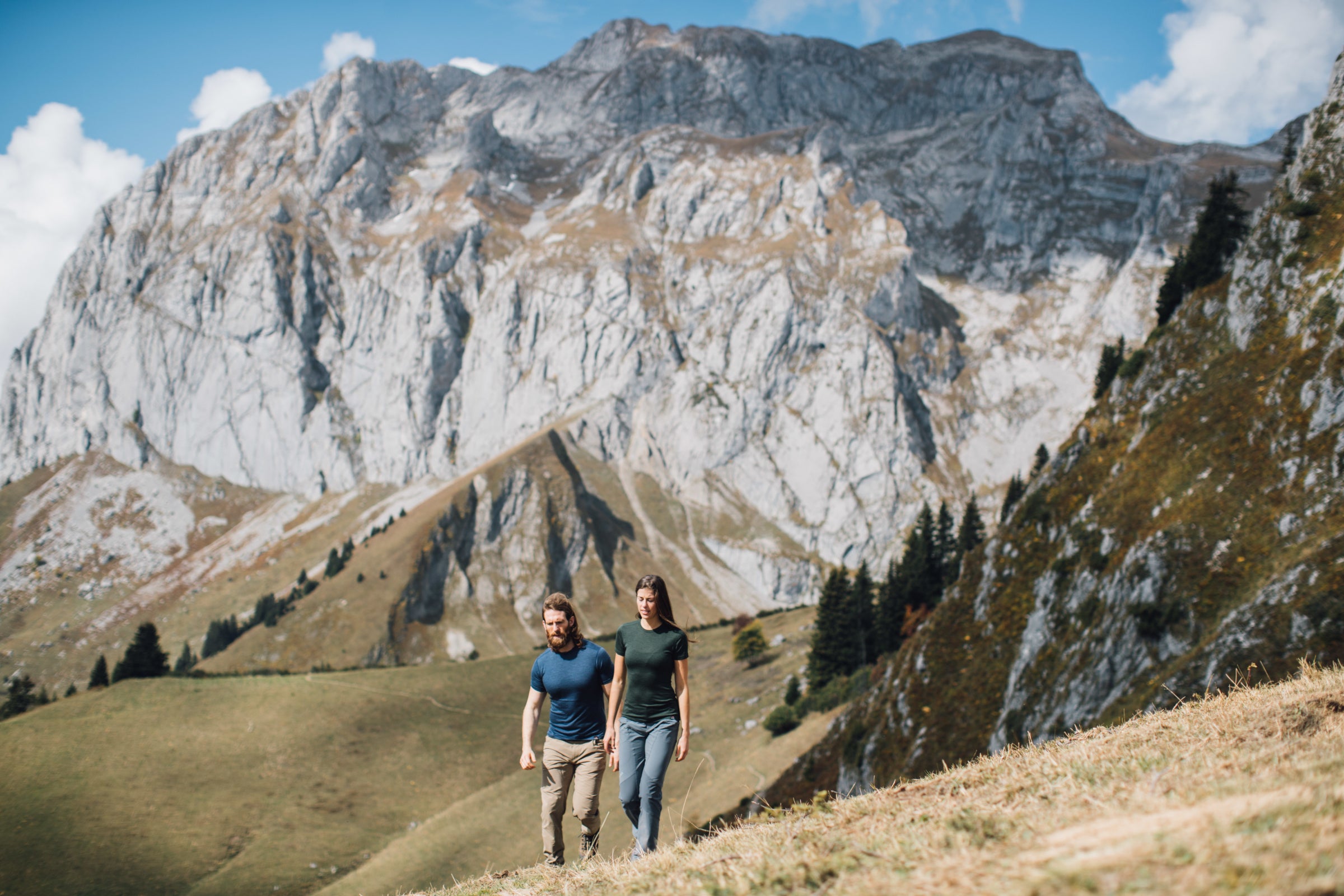 A man and a woman are hiking up a grassy slope with majestic, rocky mountains in the background, both wearing Isobaa Merino clothing