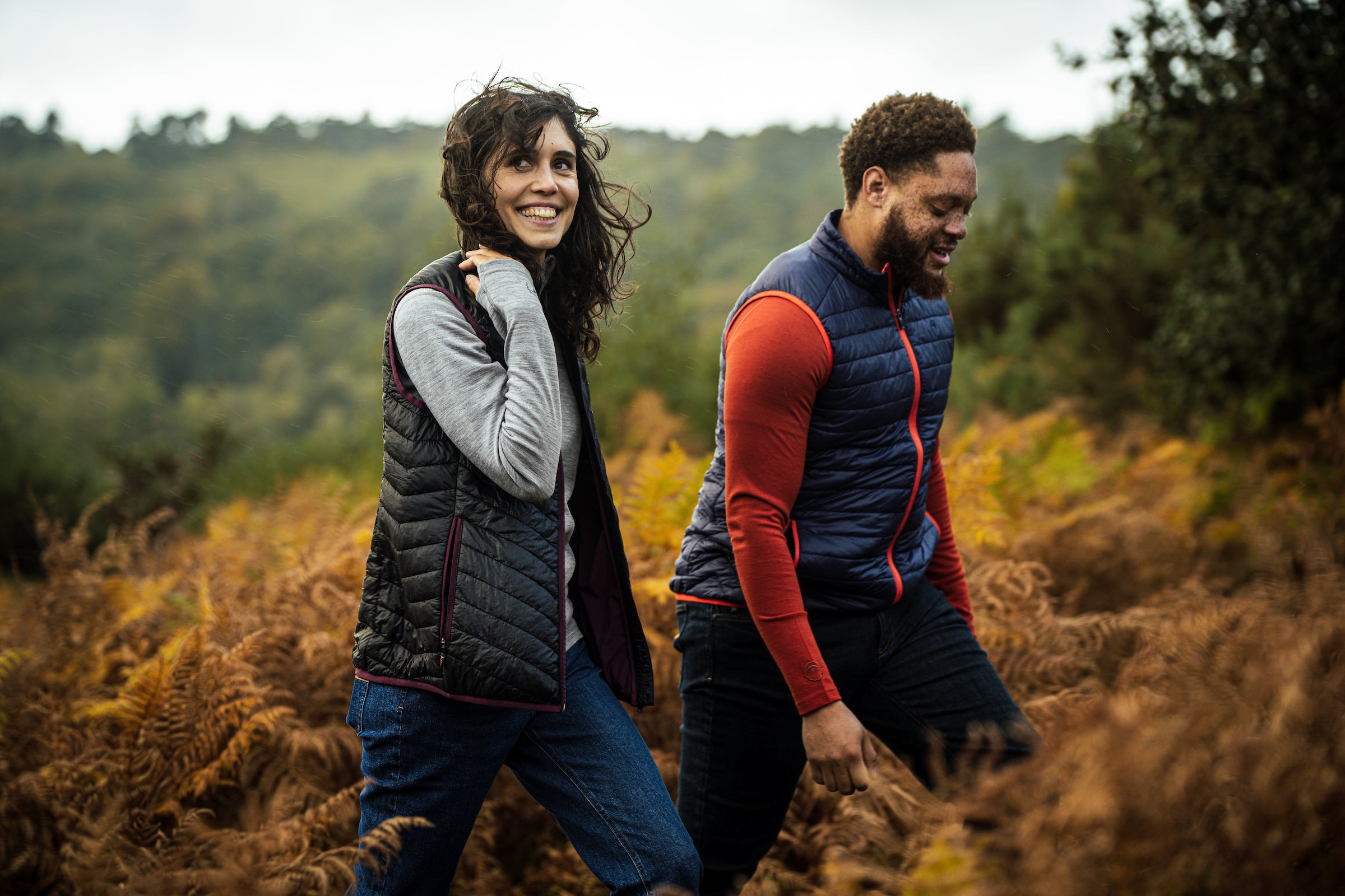 Two people walking outdoors in an autumn setting, wearing Isobaa merino wool clothing, including insulated vests and warm layers