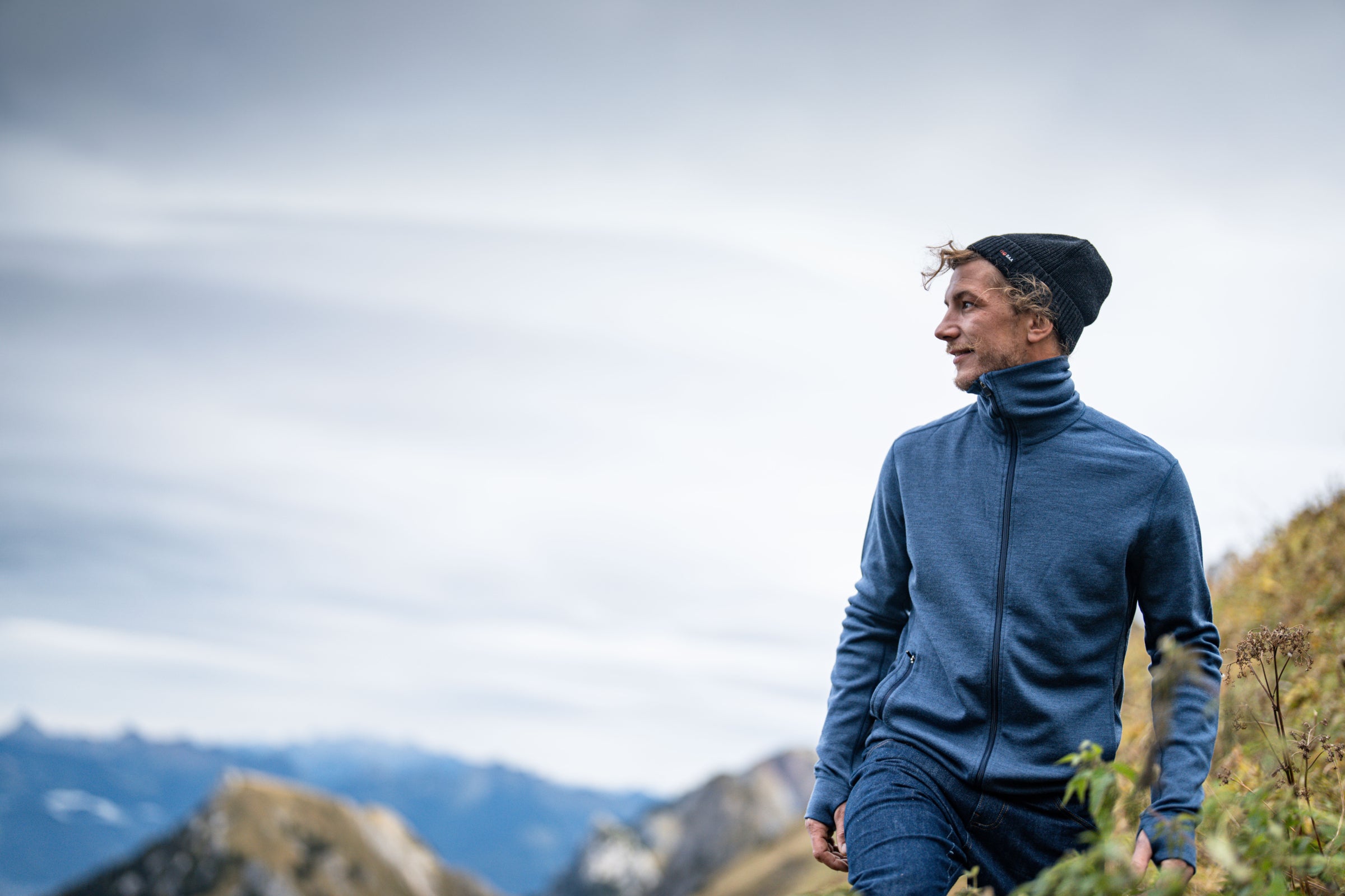 Man wearing a blue zip-up jacket and black beanie, hiking outdoors with mountains in the background