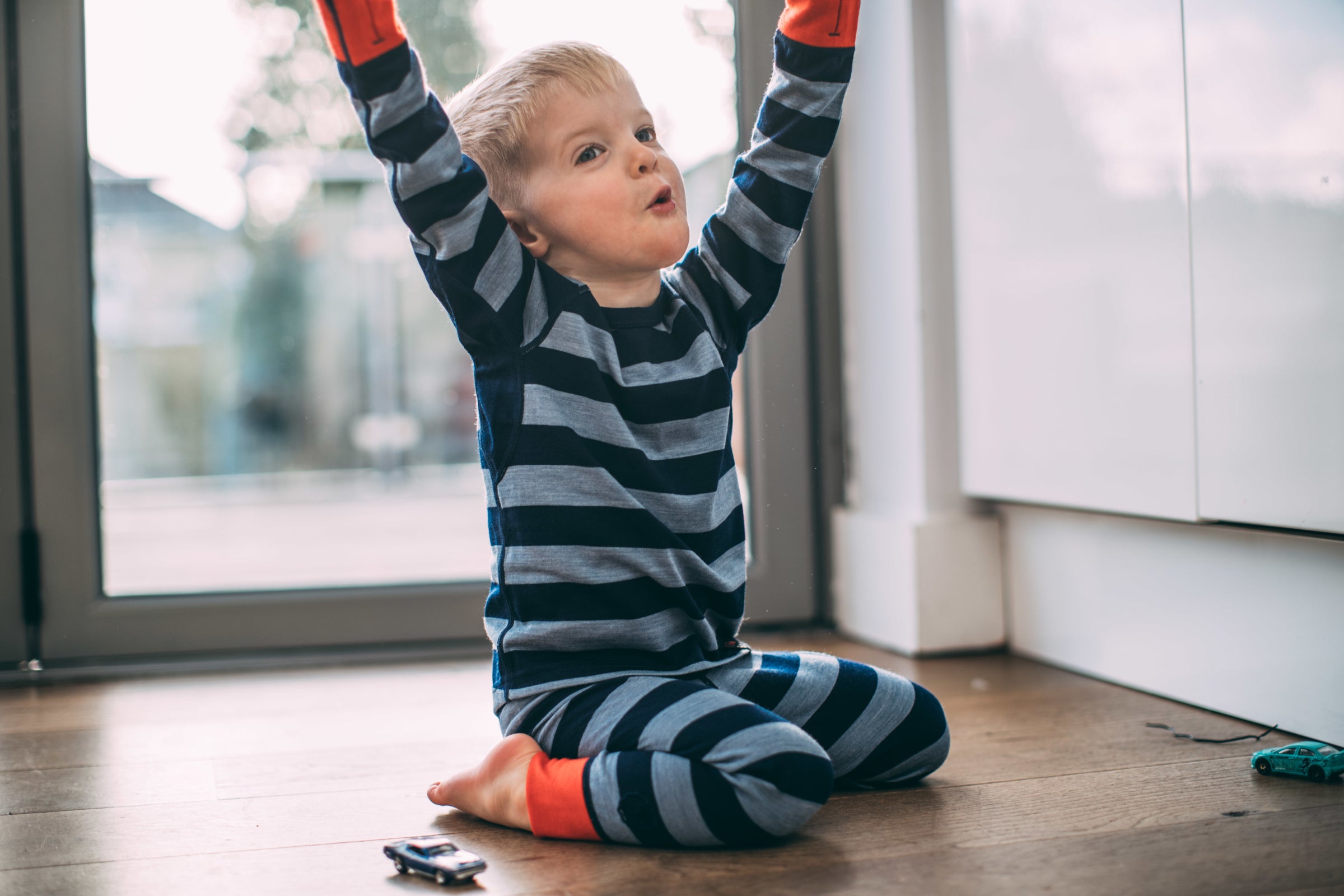 A young boy sitting on the floor indoors, raising his arms in excitement, dressed in a blue and navy striped Isobaa Merino Blend 200 Long Sleeve Crew and Merino Blend 200 Leggings