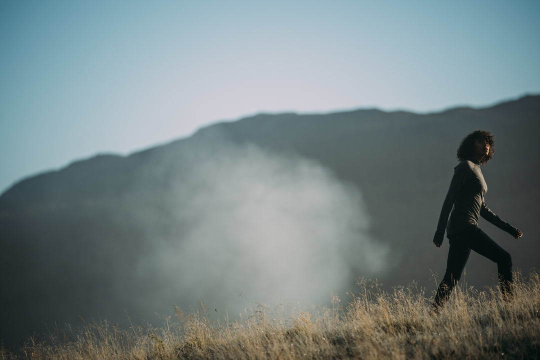 A person strides confidently across a field with tall dry grass, wearing Isobaa merino clothing, with a misty mountain backdrop and a clear sky above