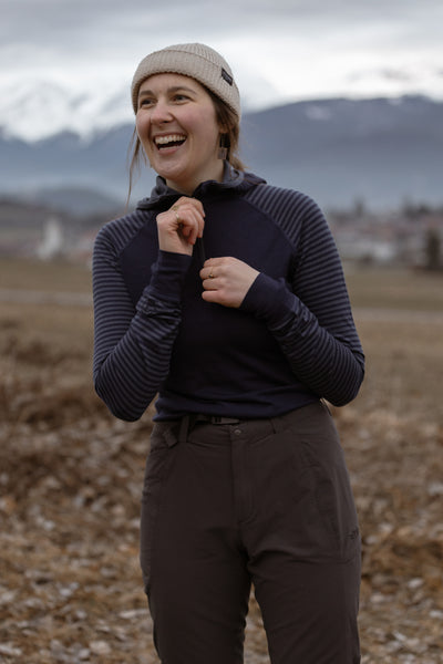 Woman in beige beanie and navy striped Isobaa merino top laughing outdoors with snowy mountains in the background