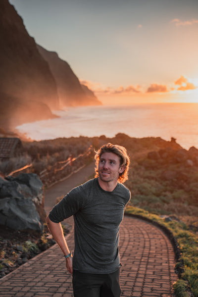 Man in grey Isobaa merino shirt standing on a coastal path at sunset with cliffs and ocean behind