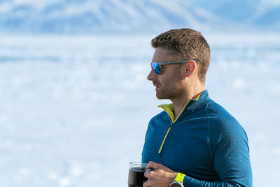 Man in petrol and lime Isobaa merino zip top holding a mug, with a snowy landscape in the background