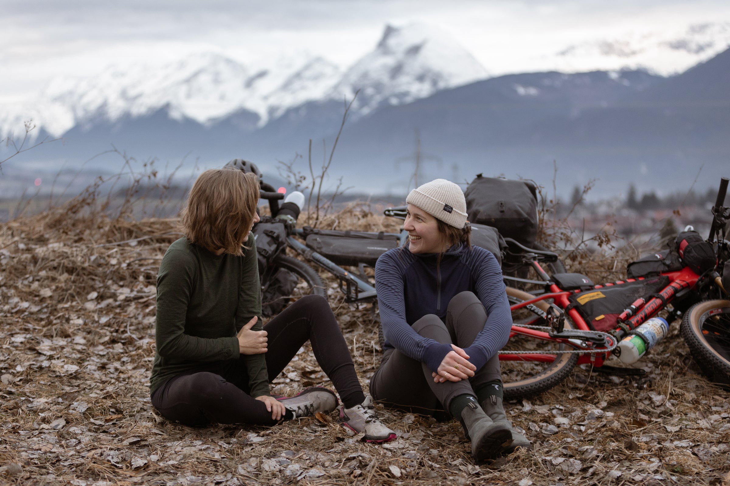 Two cyclists in merino layers resting outdoors with bikes and mountains in the background